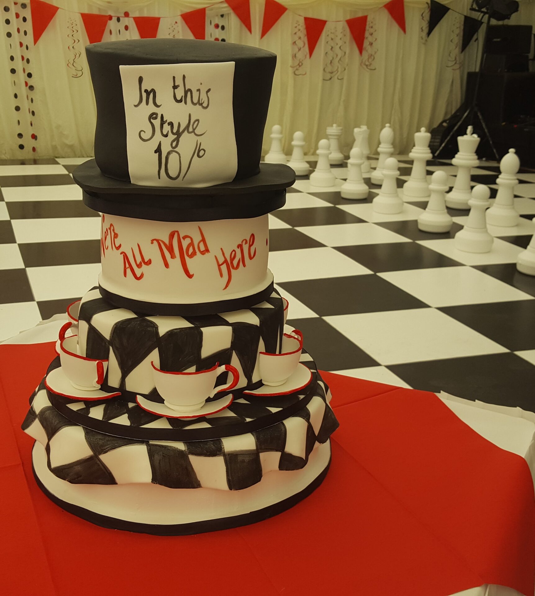 Black red and white four tier birthday cake for Alice in Wonderland themed fiftieth birthday. Bottom tier has a chequered tablecloth, next tier has same design with handmade sugar teacups stuck to the sides of the cake. Next tier has red writing "We're all mad here". Top tier is a black top hat with sales sheet "In this style". Cake is shown on a red tablecloth in front of a chess set dance floor.