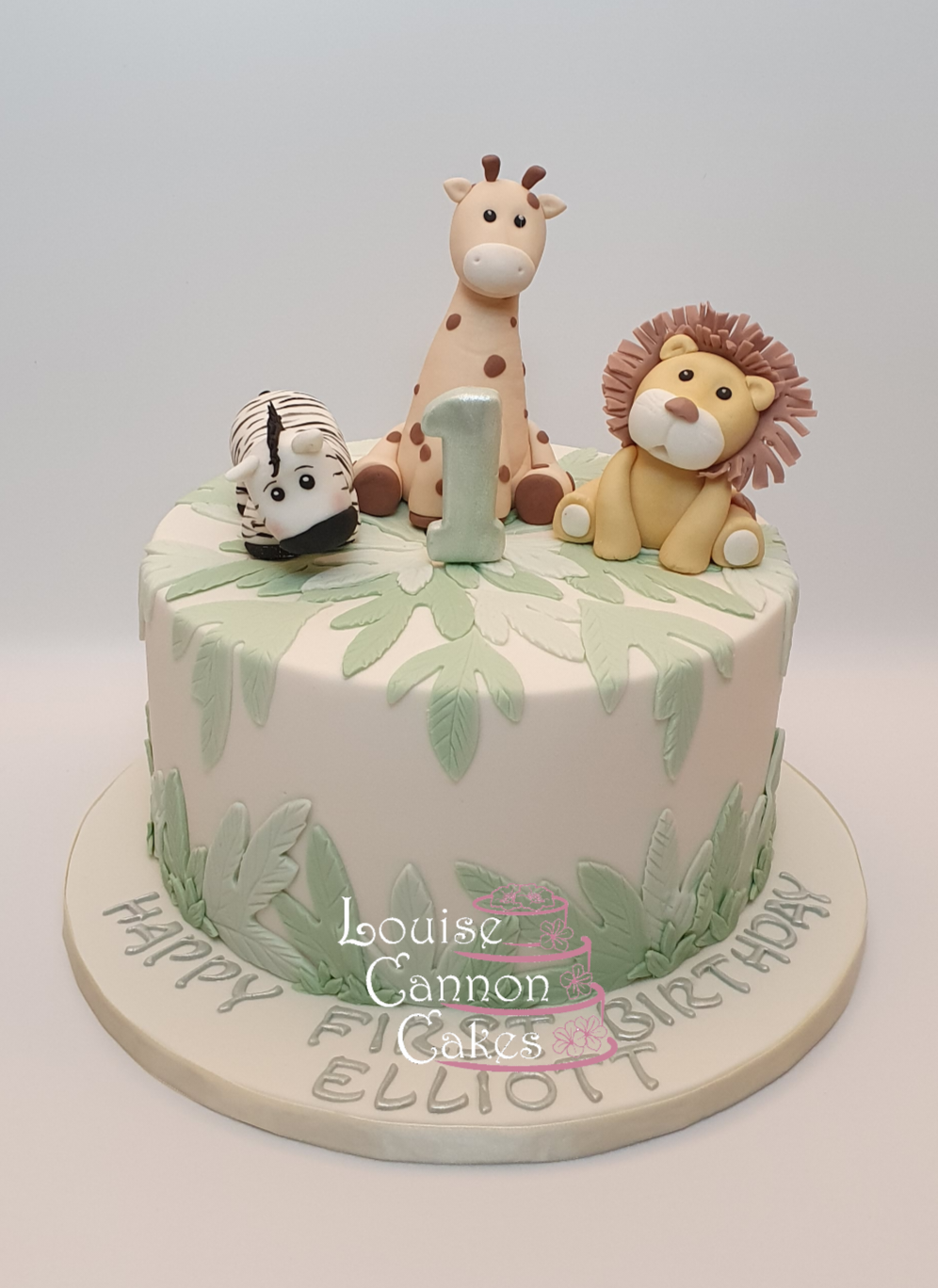 First birthday cake with jungle design. Cake is pale ivory with pale green leaves over the top and up the sides. On top are handmade fondant figures of a lion, giraffe, and zebra. Board features a happy birthday message