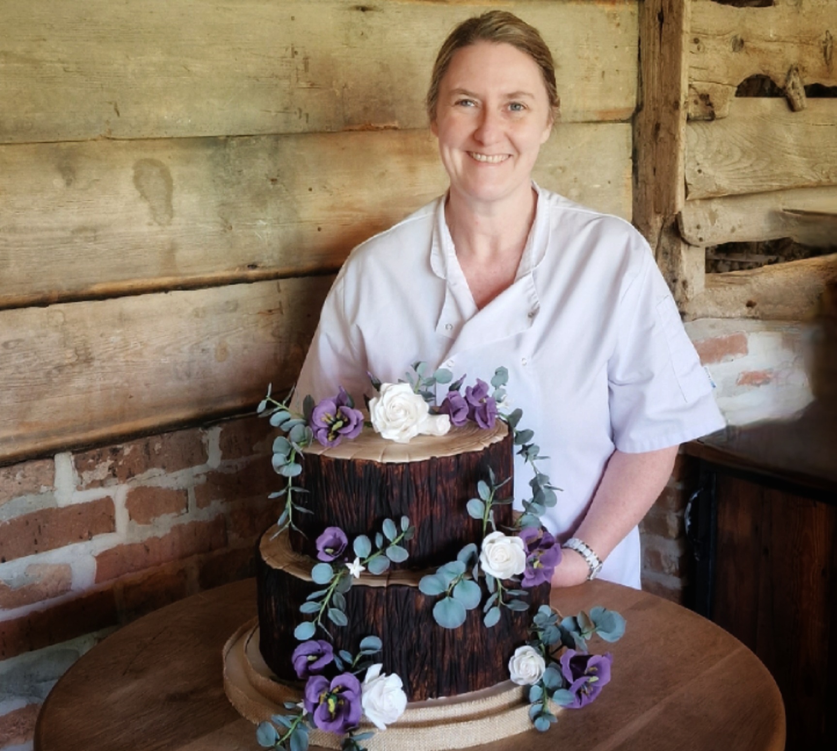 Louise with a two tier cake in the style of tree logs, with white and purple sugar flowers, and sugar eucalyptus