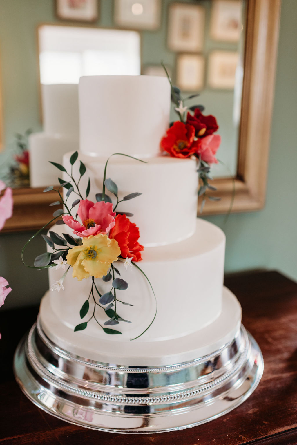 An elegant and simple white three tier wedding cake with hand-crafted red, yellow and orange sugar poppies, sugar eucalyptus and sugar ruscus leaves arranged in two sprays positioned top right and middle left Cake is set on a shiny silver cake stand, and positioned on a dark wooden sideboard in front of a large mirror hung on a pale green wall. 