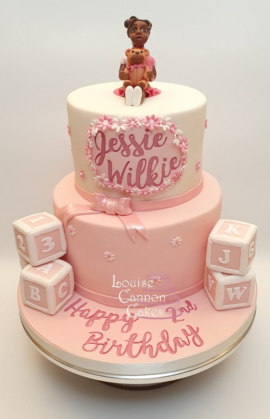 Two tier pink and white birthday cake with alphabet blocks stacked on the cake board, and a model of a girl on top hugging a teddy bear. Front of the top cake shows the girl's name, and there's a happy birthday message embossed into the pink board. 