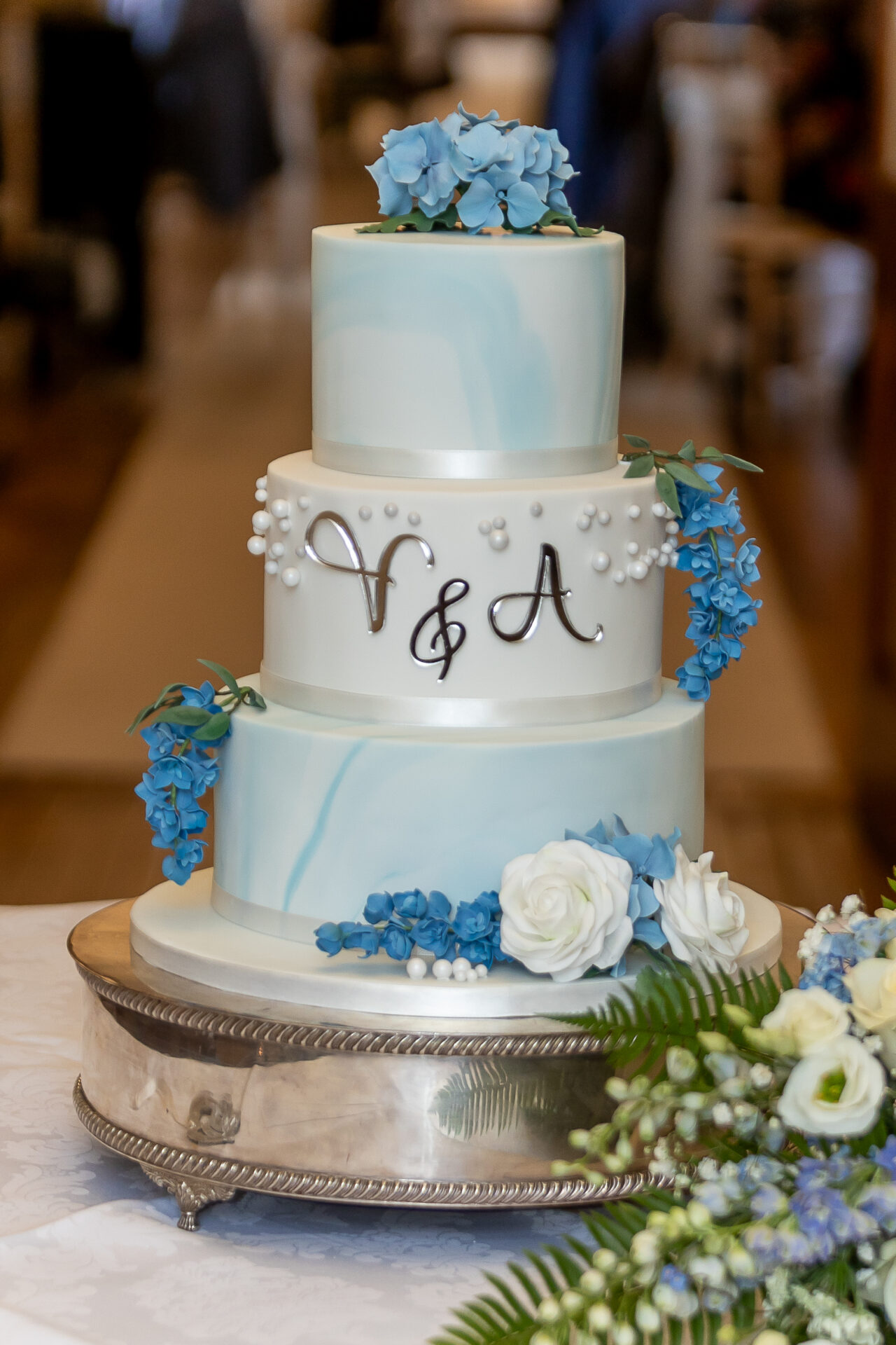 Three tier blue and white marbled cake with arrangements of handmade sugar hydrangeas, delphiniums, lisianthus and roses. Cake is displayed on a silver cake stand, set on a round table in the entrance to Clearwell Castle wedding venue. Photo taken by Stuart Harrison Photography