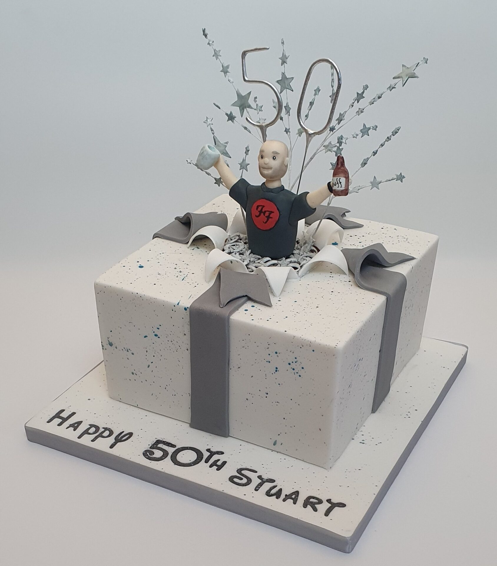 50th birthday white square cake in the shape of a wrapped present with speckled paper. With an explosion effect on top in the middle. On top of the cake is a man coming up through the "explosion" wearing a foo fighters t-shirt, and holding bottle of Duff beer in one hand an a dimple pint glass in the other. Message on board reads Happy 50th Stuart.