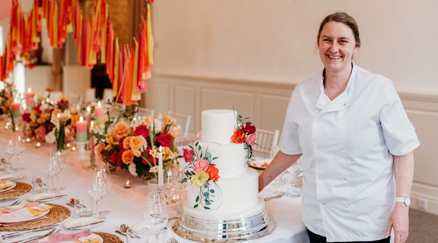 Louise Cannon (owner of Louise Cannon Cakes) stands at the end of a wedding breakfast table. She is stood next to a three tier white wedding cake with brightly coloured handmade sugar poppies and eucalyptus silver dollar stems. Picture was taken by Sarah J Photography at Merriscourt